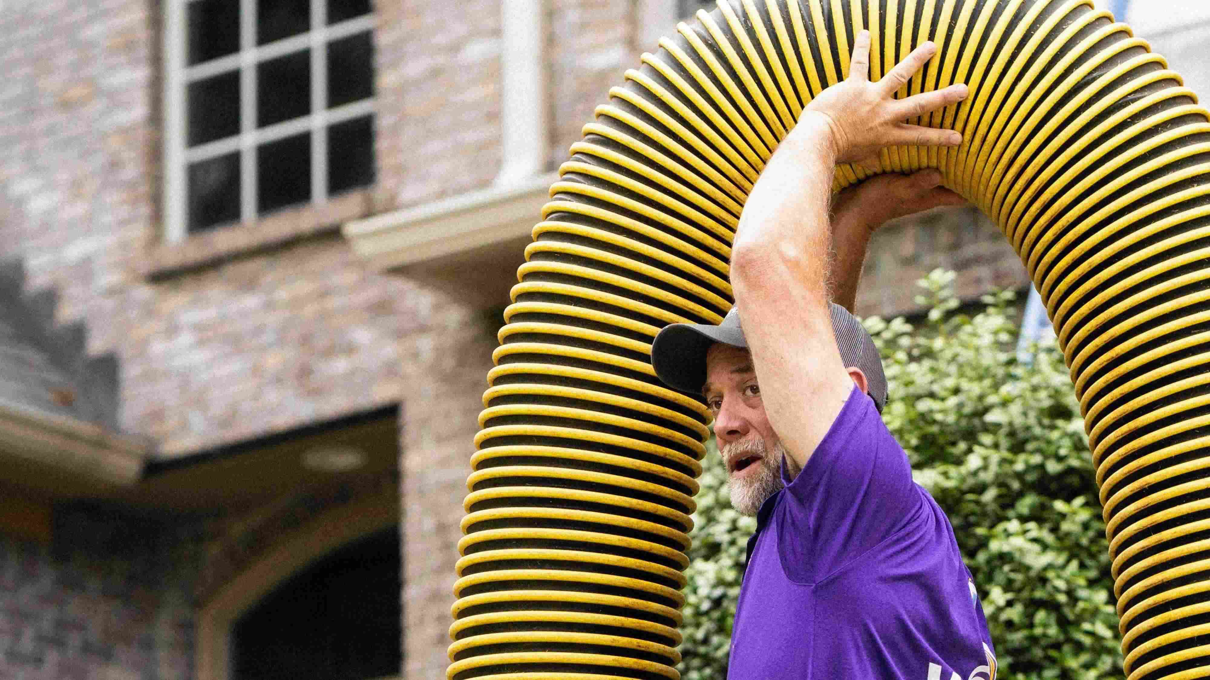 Voyager Air Duct Cleaning technician in a purple polo and cap holding a large yellow and black flexible vacuum hose arched overhead outside a brick home.